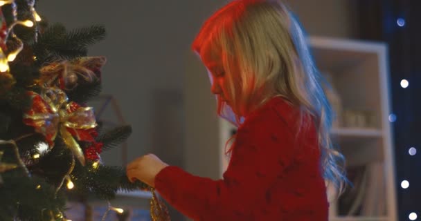 Vista laterale sulla bella e carina bambina bionda che addobba l'albero di Natale con giocattoli a casa la sera. — Video Stock