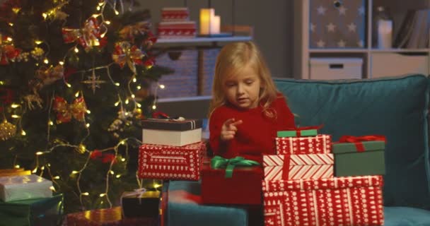 Portraitaufnahme des hübschen blonden kaukasischen Mädchens, das auf dem Sofa am Weihnachtsbaum sitzt und sich über ihr Geschenk vom Weihnachtsmann freut. — Stockvideo