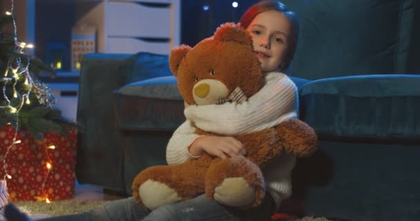 照片上的白种人漂亮的少女坐在圣诞树前的地板上，抱着大大的泰迪熊，对着摄像机微笑. — 图库视频影像