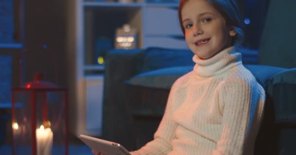 Portrait of the cute pretty small schoolgirl smiling to the camera and holding tablet device in the dark cozy room. Close up. — Stock Video