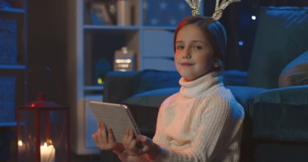 Портрет красивой маленькой девочки-подростка с забавными оленьими рогами на голове, улыбающейся в камеру при использовании планшетного компьютера вечером. Закрыть. — стоковое видео