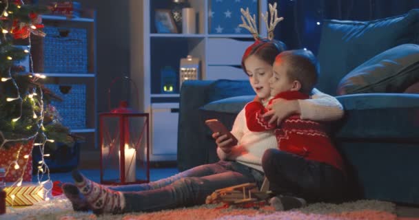 白人小孩，姐姐和弟弟，在圣诞树下的智能手机上拥抱和观看卡通片. — 图库视频影像