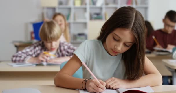 Retrato de la niña adolescente caucásica escribiendo en el copybook durante la prueba en la lección en el aula y sonriendo a la cámara. — Vídeos de Stock