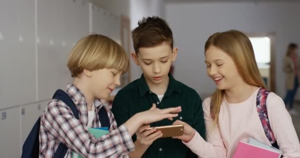 Primer plano de los alumnos caucásicos adolescentes de pie en la escuela durante el descanso y mirando el teléfono inteligente mientras juegan con interés. — Vídeo de stock