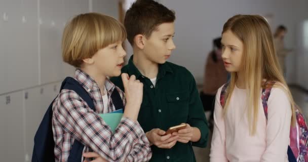 Caucasian teens friendly pupils standing at the school corridor, communicating, talking about lessons and playing on the smartphone. — Stock Video