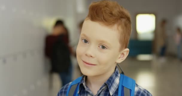 Retrato del pequeño y lindo colegial de pelo rojo caucásico con la mochila girando la cabeza hacia la cámara y sonriendo directamente a ella en el pasillo de la escuela. De cerca.. — Vídeos de Stock