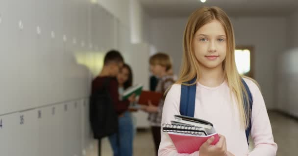 Retrato de la adolescente bonita colegiala caucásica con libros de texto y mochila de pie en el pasillo de la escuela y sonriendo a la cámara. — Vídeos de Stock