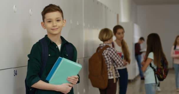 Portrait de l'adolescent écolier caucasien avec des manuels debout au passage de l'école à son étagère pendant une pause et souriant à la caméra. — Video