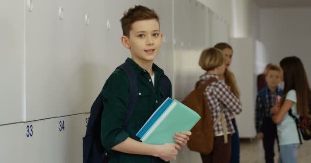 Портрет мальчика-кавказца с учебниками, поворачивающимися к камере и улыбающимися прямо к ней на школьном проходе. — стоковое видео