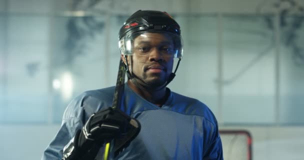 Portrait de heureux beau jeune joueur afro-américain de hockey masculin dans le casque et avec le club souriant à la caméra sur la patinoire. Gros plan de joyeux sportif attrayant en casque avec bâton. — Video