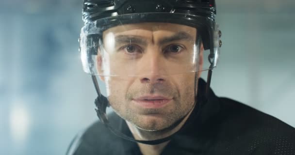 Primer plano del guapo joven jugador de hockey caucásico en casco mirando a la cámara y respirando con vapor en la arena de hielo. Retrato de deportista en casco con ojos astutos. — Vídeos de Stock