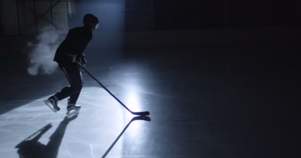 Silueta oscura del jugador de hockey masculino en uniforme, patinadores y casco deslizándose rápidamente sobre arena helada y golpeando puch con palo durante el juego. Deportista con el club golpea el disco en el hielo. — Vídeos de Stock