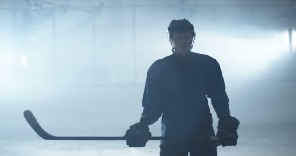 Portrait de joueur de hockey masculin en uniforme et casque debout sur une patinoire et tenant le bâton dans les mains pendant le match. Sportsman en casque avec club posant à la caméra sur glace au match. — Video