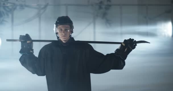 Portrait tourné sur un jeune beau joueur de hockey masculin caucasien en casque regardant la caméra et tenant le club au-dessus des épaules sur une patinoire. Sportsman en casque avec bâton. — Video