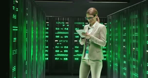 Bělošská mladá žena v brýlích stojící mezi servery s tabletovým zařízením v rukou a kontrolující velké datové procesory. Ženská analytika v ukládání dat při práci na zabezpečení digitálních informací. — Stock video