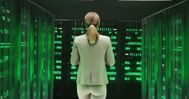 Zadní část bělošské mladé ženy s tabletem procházející mezi servery s velkými daty. Zelené pracovní procesory v úložišti databáze. Zpětný pohled na pracovnici digitální telekomunikace. — Stock video