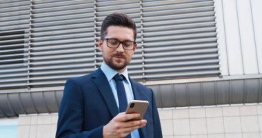 Beyaz, takım elbiseli, kravatlı, gözlüklü, akıllı telefondan mesaj atan ve dışarıda duran yakışıklı bir iş adamı. İşletme tarzında yakışıklı bir adam cep telefonuna takılıp kayıyor..