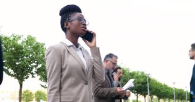Afro-Amerikalı genç iş kadını dışarıda yürüyor ve cep telefonuyla neşeli bir şekilde konuşuyor. Mutlu stil sahibi takım elbiseli güzel bir kadın işe gidiyor ve cep telefonuyla konuşuyor..