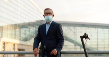 Kafkasyalı, gözlüklü ve tıbbi maskeli bir adamın elektrikli scooter 'ın önünde el ele tutuşup dik bakışının portresi. Üst düzey erkek, solunum koruması altında.
