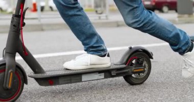 Elektrikli scooter 'ın üzerinde duran beyaz spor ayakkabılı erkek ayaklarının yakınına. Şehir caddesinde sürüklenen bir yol. Kasabada hızlı giden bir adam. Modern araç. Şehir ulaşımı.