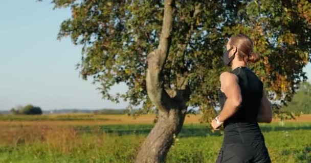 Traseiro em branco bonito forte corredor masculino correndo na natureza pela manhã no dia de verão. Um desportista atraente mascarado a correr lá fora. Conceito pandêmico. Visão traseira. — Vídeo de Stock