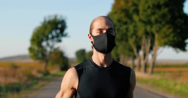 Gros plan du beau caucasien fort jogger masculin avec les muscles en cours d'exécution sur la route dans la campagne le jour d'été. Attrayant homme sportif en masque jogging à l'extérieur. Concept pandémique. — Video