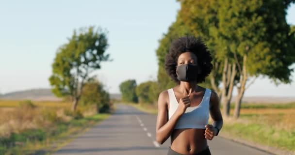 Africano americano bonito slim feminino corredor com cabelo encaracolado e no topo correndo na estrada no campo no dia de verão. Mulher desportiva atraente com máscara a correr lá fora. Conceito de pandemia. — Vídeo de Stock