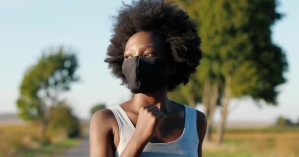 Närbild av afroamerikanska vackra kvinnliga joggare med lockigt hår körs på väg på landsbygden på sommardagen på morgonen. Ganska sportig kvinna i mask joggar utanför. Begreppet coronavirus. — Stockvideo