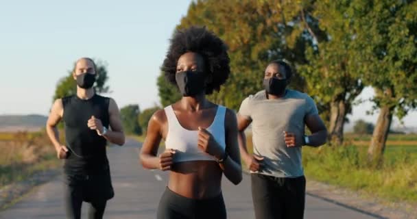 Mischlingshündchen und Joggerinnen laufen im Sommer auf der Landstraße. Afroamerikaner mit schlanker, sportlicher Frau und multiethnischen Männern beim Joggen im Freien. Läufer in Masken bei Pandemie. — Stockvideo