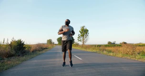 Afroamerikaner, gut aussehender starker männlicher Jogger, der an Sommertagen auf dem Land unterwegs ist. Attraktiver, sportlicher Mann mit Maske, der draußen joggt. Pandemisches Coronavirus-Konzept. Sportler. — Stockvideo