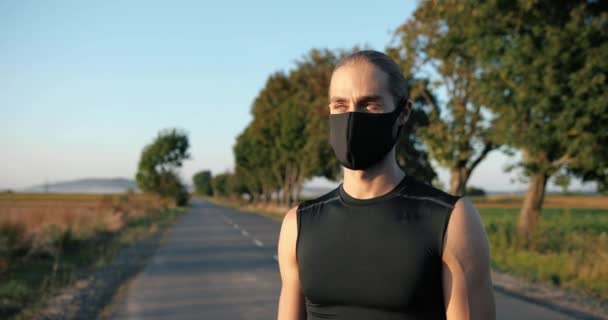 Портрет сильного красивого молодого кавказского спортсмена в маске, стоящего на дороге в сельской местности и смотрящего в камеру. Привлекательный спортивный бегун в летний день. Долли выстрелила. Увеличение в лицо. Закрыть. — стоковое видео