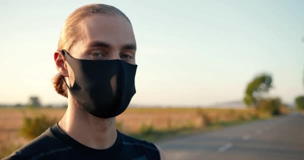 Gros plan d'un beau jeune sportif caucasien masqué debout sur la route dans la campagne et se tournant face à la caméra. Portrait d'attrayant sportif gars jogger le jour de l'été. — Video