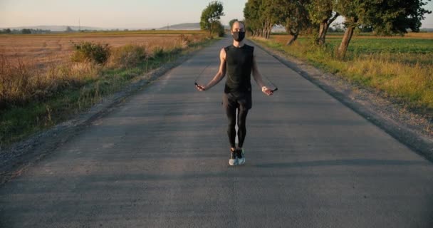 Кавказский красивый молодой спортсмен бегун в маске прыгает по скакалке по сельской дороге на природе в летний день. Спортивный бегун на скакалке под открытым небом. — стоковое видео
