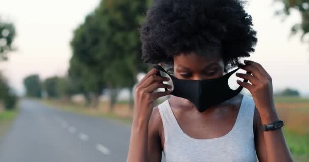 Primer plano de la joven afroamericana deportista con el pelo rizado tomando la máscara en la boca durante la pandemia de corona mientras está de pie en el camino del campo. Corredora preparándose para correr. — Vídeo de stock