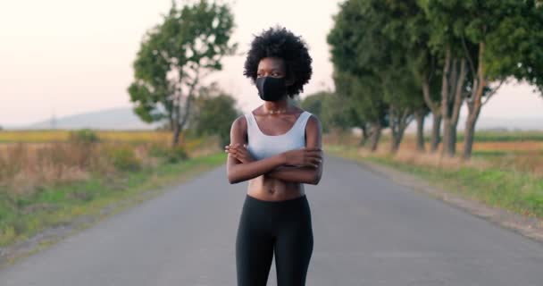 Portrait of slim beautiful young African American sportswoman runner in mask standing at road in countryside, crossing hands and looking at camera. Sporty woman jogger on summer day. Pandemic concept. — Stock Video