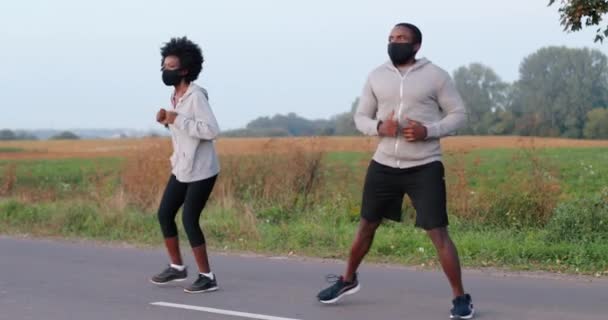 Afroamerikanska unga par joggare i masker som tränar tillsammans i naturen på sommardagen. Sportman och idrottskvinna joggar på landsbygden under coronavirus. Manliga och kvinnliga löpare värmer upp. — Stockvideo