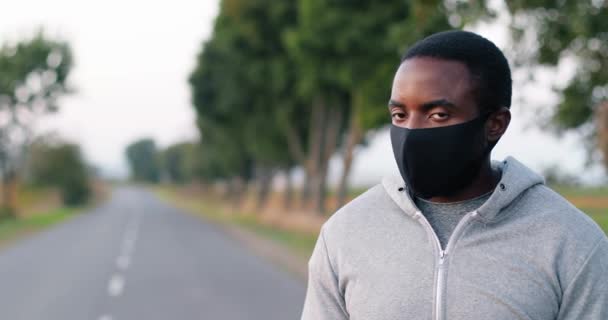 Portret van een knappe jonge Afro-Amerikaanse sportman met masker op het platteland die gezicht naar camera draait. Sluiten van sportieve kerel jogger staan op de weg op zomerdag. Runner tijdens pandemie. — Stockvideo