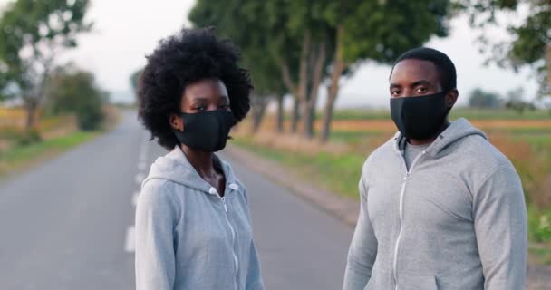 Retrato de una joven pareja afroamericana de corredores con máscaras paradas juntas en la naturaleza en el día de verano. Deportista y deportista trota en la carretera rural durante la pandemia. Corredores masculinos y femeninos. — Vídeos de Stock