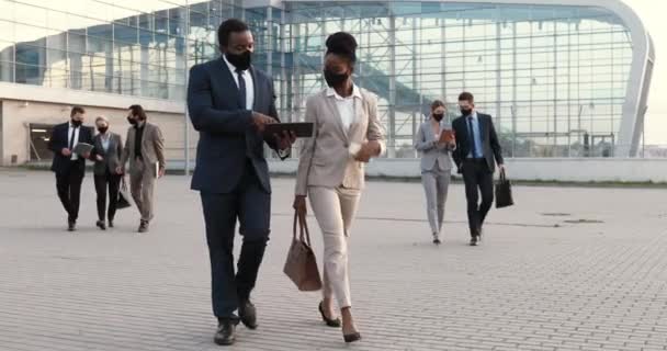 Afro-Amerikaanse jonge paar zakenpartners in maskers lopen naar het werk, praten en het gebruik van tablet apparaat buiten in de financiële stad. Zakenman en zakenvrouw in gesprek op straat. — Stockvideo