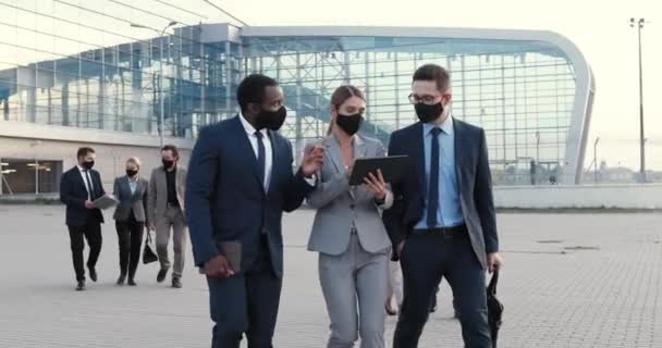 Kaukasische zakenvrouw en twee gemengde rassen zakenmannen in maskers lopen buiten in het business center en praten met tablet apparaat in handen. Multi-etnische werkpartners bespreken project en financiering. — Stockvideo