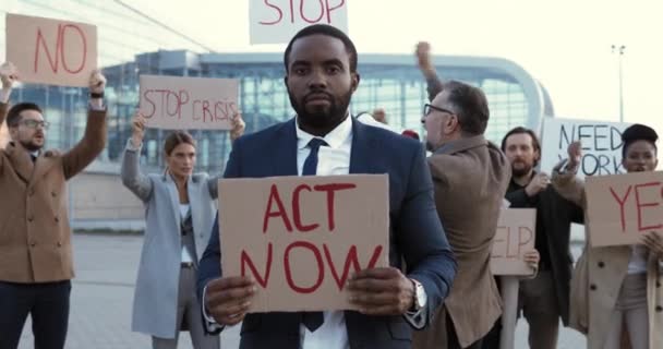 Portrait de l'homme afro-américain en costume et cravate debout à l'extérieur pour protester avec affiche Act Now. Manifestant masculin en grève contre le chômage. Des gens métis en arrière-plan. Manifestation. Frappe. — Video