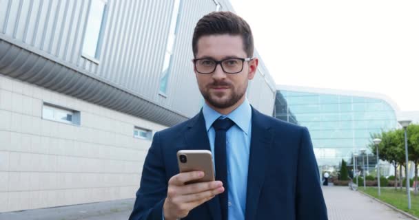 Retrato de joven hombre de negocios guapo caucásico en traje, corbata y gafas enviando mensajes de texto en el teléfono inteligente y sonriendo a la cámara al aire libre. Hombre guapo en estilo de negocio tocando y desplazándose en el teléfono móvil — Vídeos de Stock