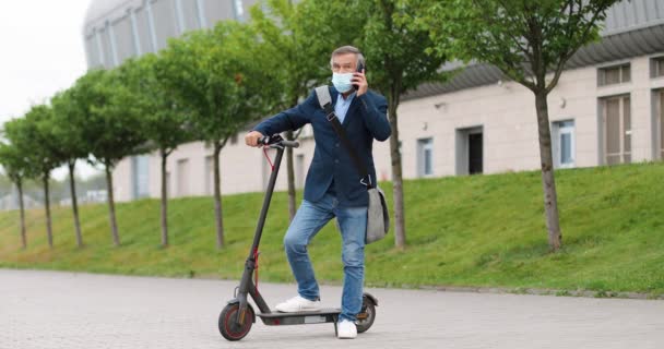 Старший седой мужчина в медицинской маске стоит на электрическом скутере на улице в городе и разговаривает по мобильному телефону. Старый дед говорит по телефону на улице. Звонок. Разговор. — стоковое видео