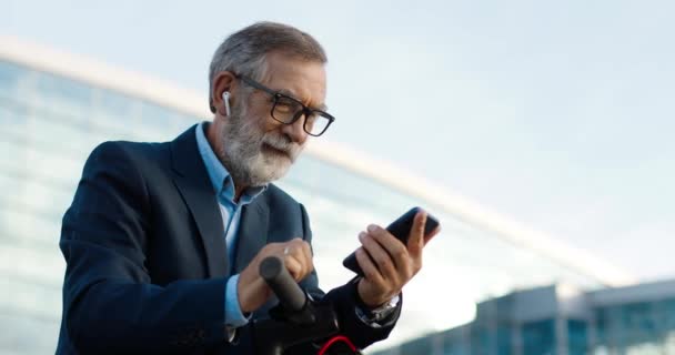 Senior gråhårig man i glasögon och hörlurar stående på cykel på gatan och knacka eller rulla på mobiltelefon. Gammal farfar i airpods med smartphone och lyssna på musik utomhus. — Stockvideo