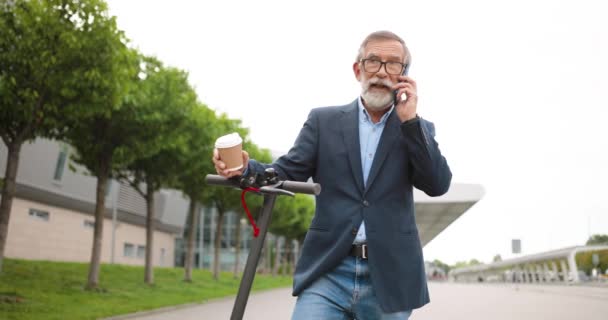 Hombre mayor con gafas parado en scooter eléctrico en la ciudad, sosteniendo café para llevar y hablando por teléfono móvil. Viejo abuelo hablando por celular al aire libre. Llamada en la calle. Conversación. — Vídeos de Stock