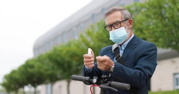 Hombre mayor de pelo gris con gafas y máscara médica parado en bicicleta en la calle y desinfectando las manos con desinfectante en aerosol. Viejo abuelo pulverización desinfección y limpieza de la piel de los gérmenes al aire libre. — Vídeos de Stock