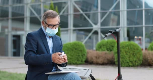 戴眼镜和医疗面具的老人坐在街上的电动车旁，用喷雾清洁剂消毒双手。老爷爷在膝上型计算机工作前喷洒细菌消毒. — 图库视频影像