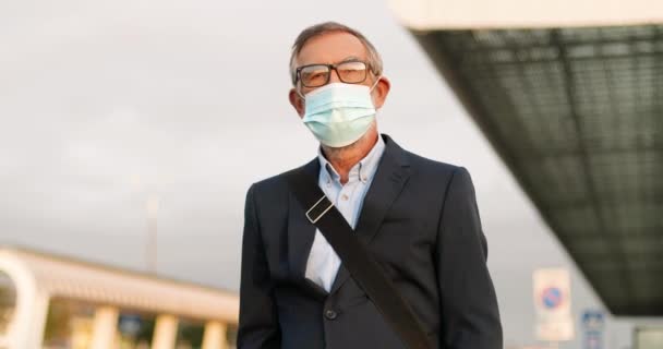 Portret van een knappe blanke oude man met een bril en een medisch masker die in de buitenlucht staat in een stedelijke omgeving en naar de camera kijkt. Oudere grijze man in bril en ademhalingsbescherming. — Stockvideo