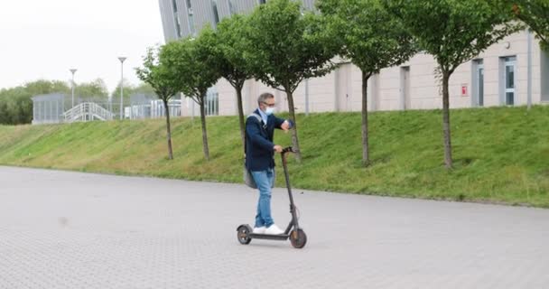 Viejo hombre de pelo gris con anteojos y máscara médica de pie en scooter eléctrico y montar en la calle de la ciudad. Hombre mayor corriendo a alguna parte y mirando el reloj al aire libre. Concepto de vehículo urbano. — Vídeo de stock