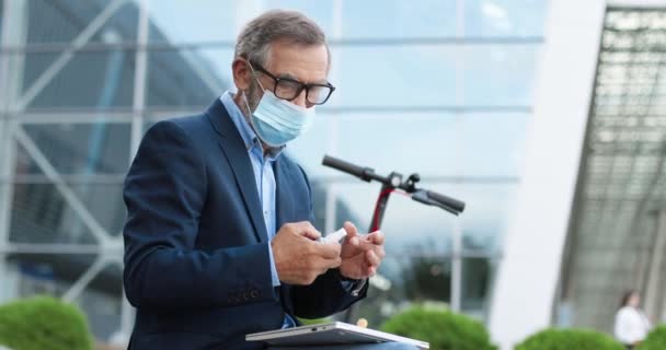 Ανώτερος άνθρωπος με γυαλιά και ιατρική μάσκα κάθεται σε ηλεκτρικά σκούτερ στο δρόμο και απολυμαίνοντας τα χέρια με απολυμαντικό σπρέι. Παλιός επιχειρηματίας ψεκάζει απολύμανση από μικρόβια πριν από την εργασία υπολογιστή laptop. — Αρχείο Βίντεο